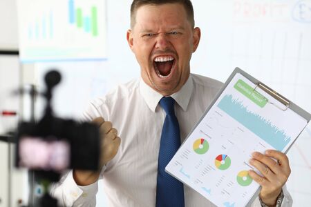 Emotions head company from statistics report. Man shoots video on professional camera. He screams, clenched one hand in fist, and in other hold tablet with documents. In background office room.の写真素材