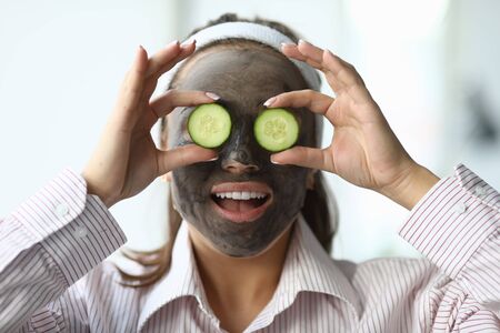 Cosmetic procedure for cleansing, refreshing skin. Girl made black wellness mask, holds in hands slices cucumber and applies them to her eyes. She put on special hair bandage and smiles.の写真素材