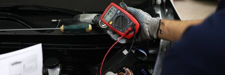 Close-up of male hands checking voltage of automobile battery under hood of automobile with accuracy. Mechanic holding modern utility. Automotive checkup conceptの写真素材