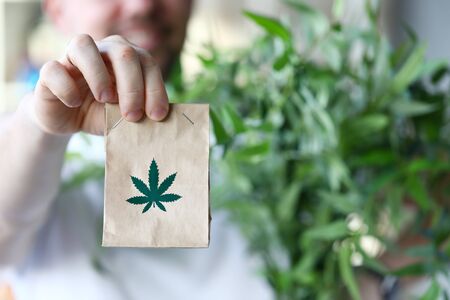 Male hand hold paper packet with marijuana symbol closeup background. Online shopping sativa concept