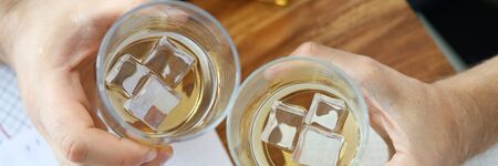 Top view on employee hands holding two glass of whiskey. Alcohol beverage with ice cubes. Workers sitting at table with papers and drink, relaxation in officeの写真素材