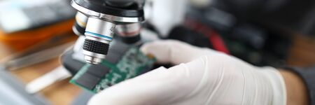 Close-up of laptop hardware board fixing at pc service center. Male hands holding circuit. Specialist looking at computer part through microscope. Detailed analysis of gadgets component productivityの写真素材