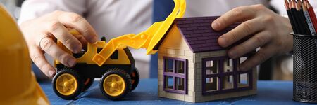 Close-up of male hands with yellow toy excavator. Builder driving backhoe loader on blueprint. Businessman in white shirt with tie. Construction and business conceptの写真素材