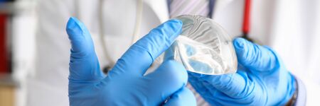 Male doctor hand in blue protective gloves hold new fashion silicone breast implant closeup. Plastic...