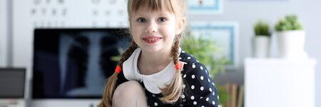 Portrait of cheerful girl in polka-dot dress sitting in doctor office. Joyful kid visiting ophthalmologist and looking at camera with happiness and gladness. Healthy lifestyle conceptの写真素材