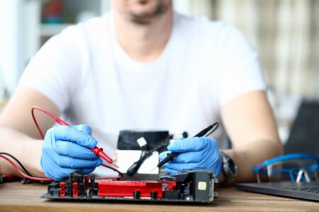 Close-up of male wearing protective blue gloves and using voltmeter on repairing detail. Middle-aged...