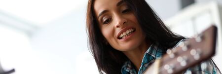 Portrait of attractive smiling woman learning play guitar and looking at strings carefully. Charming...
