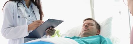 Portrait of beautiful doctor asking upset wife about well-being ill husband. Unhealthy patient dripping dropper in hospital ward. Healthcare and treatments conceptの写真素材