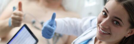 Portrait of cheerful doc standing in modern hospital ward and looking at camera with happiness. Ill man lying on medical couch and showing approving thumb up gesture with therapist. Hospital conceptの写真素材