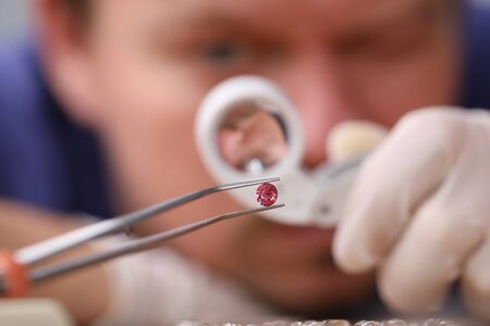 Male looks through magnifying glass brink diamond. Man checks safety product and stone, cleans and polishes it if necessary. Regular inspection jewelry to identify possible problems or damageの写真素材