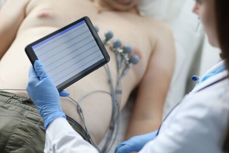 Close-up of female doctor cardiologist check patient results on modern tablet. Protective uniform and gloves. Physician woman diagnostic mans heart. Medicine and ecg conceptの写真素材