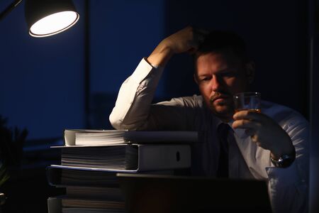 Portrait of male relaxing after intense workday with pile of paper folders full of corporation documents. Tired businessman sitting indoors and looking at alcohol in glassful. Night at office conceptの写真素材