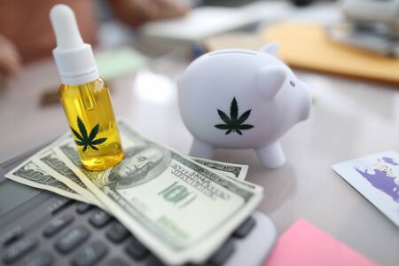 Close-up of white piggy bank with marijuana sign, cannabinoid oil in bottle with pipette. Cash on table. Person paying for pain medication. Painkillers conceptの写真素材