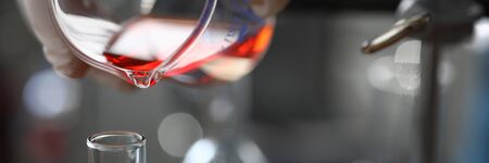 Close-up of assistant hand holding flask. Chemist pouring red liquid into vial. Researcher making mixture of fluid for experiment in lab. Chemical experiments conceptの写真素材