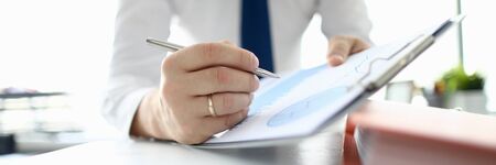 Close-up of businessman hand signing important documents. Manager making annual report and writing down on clipboard. Man in suit sitting in company office. Business and finance conceptの写真素材