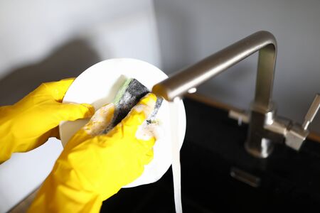 Close-up of person washing dishes under water. Housewife clean plate. Kitchen silver sink and green sponge. Man wearing yellow protective gloves. Housework conceptの写真素材