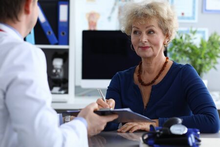 Portrait of elderly woman at doctors appointment. Female signing prescription in medical worker cabinet. Blood pressure on medic table. Medicine and qualified specialist conceptの写真素材