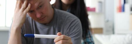 Portrait of anxious worried man holding pregnancy test in hand. Troublous female sitting behind and supporting male. People in cozy bedroom. Unexpected pregnancy conceptの写真素材