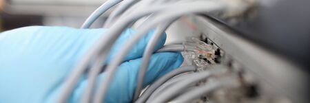 Close-up view of persons hand holding grey cable. Network panel with lots of wires. Power unit and...