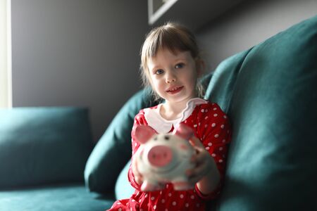 Portrait of smiling beautiful child holding piggy bank sitting on sofa indoors. Lovely blonde kid...