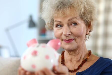 Close-up of attractive elderly woman holding piggy bank for money. Colourful container for coins and cash. Beautiful female indoors. Saving for future and economy conceptの写真素材
