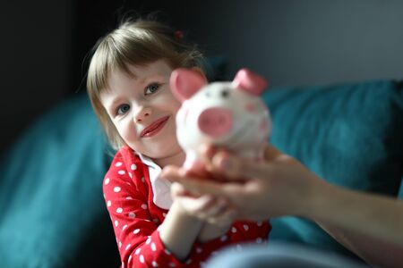Portrait of lovely little kid holding pink piggy bank. Smiling light haired girl wearing red dress in dots. Charming toddler posing on camera. Money and saving up for toy conceptの写真素材