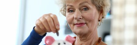 Elderly woman happily saves money in piggy bank. Older generation is used to saving and saving money. Closeup, an elderly woman is happy with her savings and puts coin in piggy bank.の写真素材