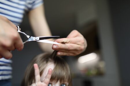 Close-up of mother holding strand of daughter hair and going to cut with metal scissors. Girl showing victory sign. Hairdresser at home during lockdown. Family time together conceptの写真素材