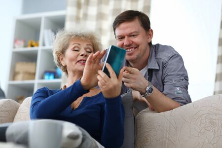 Portrait of cheerful son and mother watching old photos on smartphone. Smiling elderly woman and middle-aged man spending time at home. Precious memory ad leisure conceptの写真素材