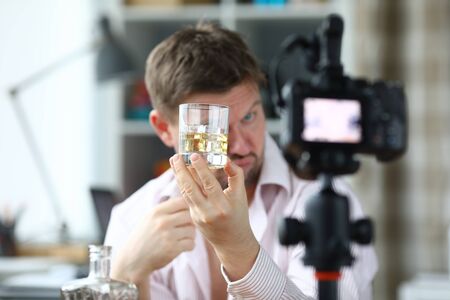Drunk man looks glass alcohol and takes off camera. Guy drinks at home during quarantine. Video blogger tastes alcoholic drinks for subscribers. Falsification branded alcohol videoの写真素材