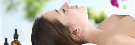 Beauty treatments in salon using organic oil. Beautiful girl lies on table for cosmetic procedures, under her head roller from towel, next to glass jars with massage oil. Pleasant sensations.の写真素材