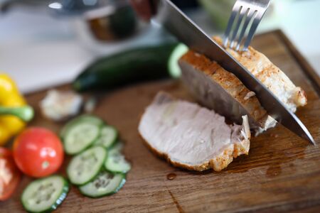 Close-up of person cutting juicy slice of baked meat with sharp knife and fork. Pieces of cucumber and tomato on wooden board. Diet nutrition. Summer healthy food conceptの写真素材