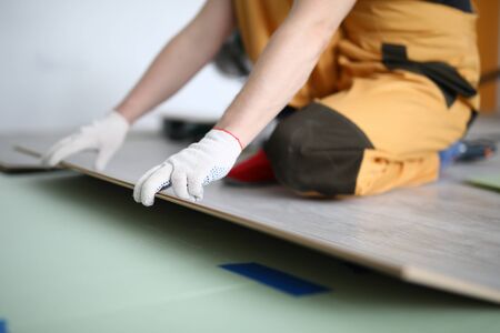 Close-up of adult man installing new laminated wooden floor in room indoors. Professional qualified worker in protective white gloves. Foreman in yellow costume. Construction site conceptの写真素材
