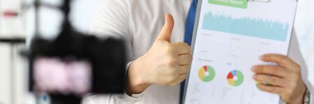 Focus on male hand showing thumb up sign for camera. Man standing in office demonstrating statistics documents with charts, graphs and diagram. Guy making video for vlogの写真素材