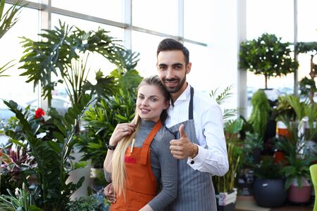 Joyful bearded man in apron hugging smiling young woman and giving thumbs up while standing in plant storeの写真素材