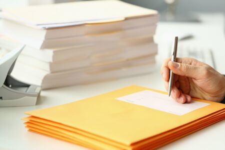 Close up of young man with pen in hand signing envelope while sitting at the table with documents