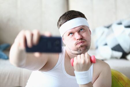 Young man in sport headband doing exercise with dumbbell and trying not to breath while taking picture with smartphoneの写真素材