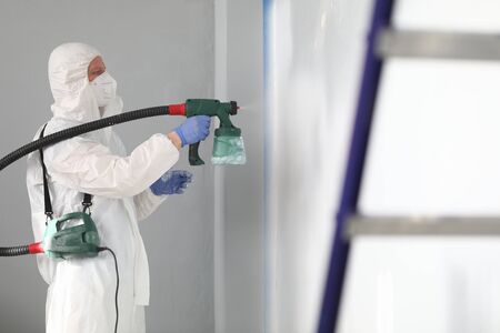 Close-up of professional male worker painting wall in grey colour with spray gun. Adult man in white protective costume and face mask. Black metal ladder in right side. Renovation conceptの写真素材