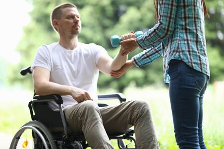 Close-up of middle-aged man sitting in wheelchair and trying to exercise with blue dumbbell. Woman helping male training arm. Sport and active lifestyle. Disabled people conceptの写真素材