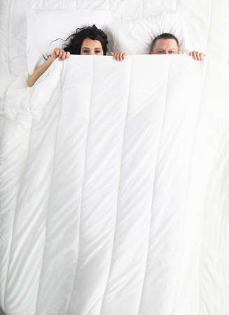 Top view of man and woman hiding under duvet. Happy funny couple laying together in bed. Lovely relationship in family. White bedding. Wake up and good morning conceptの写真素材