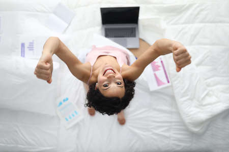 Top view of happy young woman sitting in bed. Active and cheerful female clench fists. Beautiful lady working from home. Laptop on white sheets. Job and technology conceptの写真素材