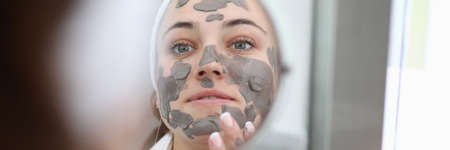 Close-up view of beautiful young woman applying clay mask on face. Female looking in small mirror. Moisturizing treatment. Morning ritual and skincare procedure conceptの写真素材