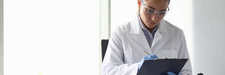 Portrait of concentrated woman looking at documents with seriousness. Lab assistant writing results of science experiment. Chemical investigations conceptの写真素材