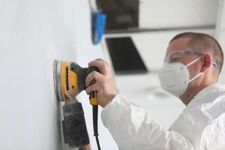 Close-up of professional worker wearing white protective face mask and working with yellow sander...