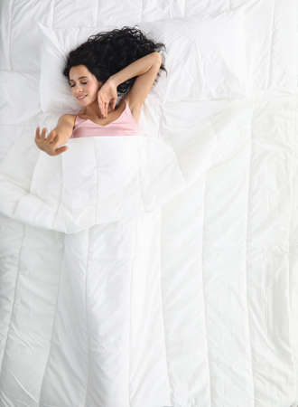 Top view of stretching young woman resting in bed. Charming female with brunette hair down. Well rested lady after night sleep. Good morning and new day conceptの写真素材