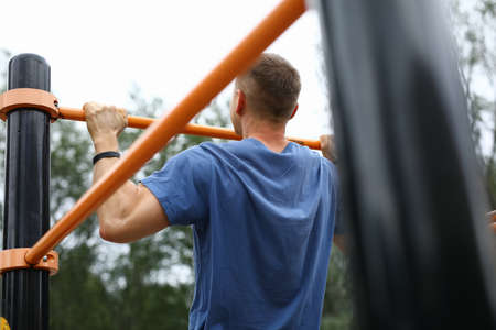 Close-up of strong male pulls up on street horizontal bar. Person in blue shirt training outdoors....