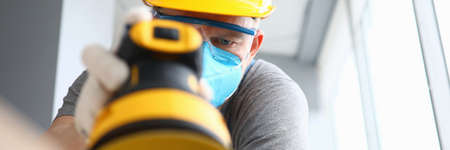 Close-up of construction worker using sander machine wearing protective yellow helmet and gloves. Repairman polishing wood in apartment. Renovation conceptの写真素材