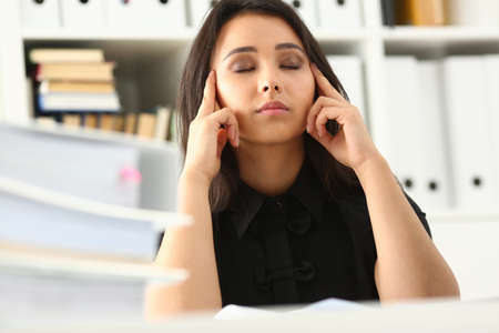 Exhausted of paper work in office female is sitting at desk and closing eyes while holding fingers...