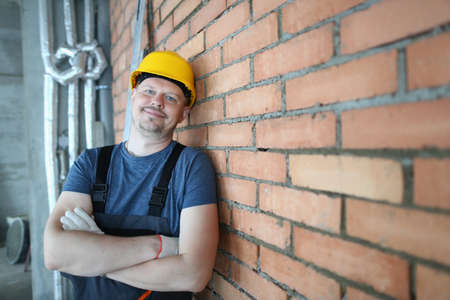 Waist up portrait of smiling bearded man in yellow hard hat standing and leaning against brick wall