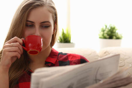 Relaxed young female person being at home and reading interesting newspaper, enjoying her favorite drinkの写真素材